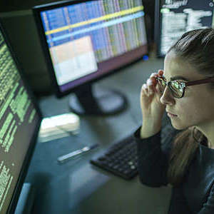 A young woman is seated at a desk surrounded by monitors displaying data.