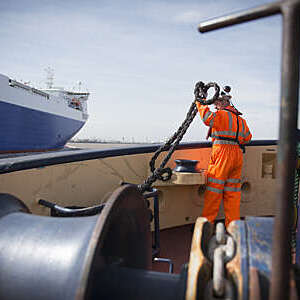 Tug worker in protective orange clothing holding a rope on deck.