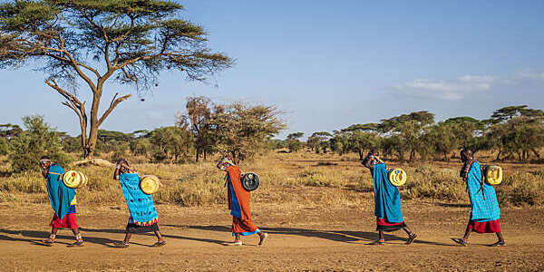 Five African women walk across the barren landscape carrying water tanks on their backs.